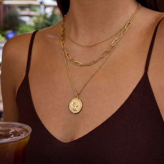 A close-up of a woman wearing layered gold necklaces, including a coin pendant necklace, a paperclip chain, and a delicate chain, paired with a dark brown ribbed camisole top.