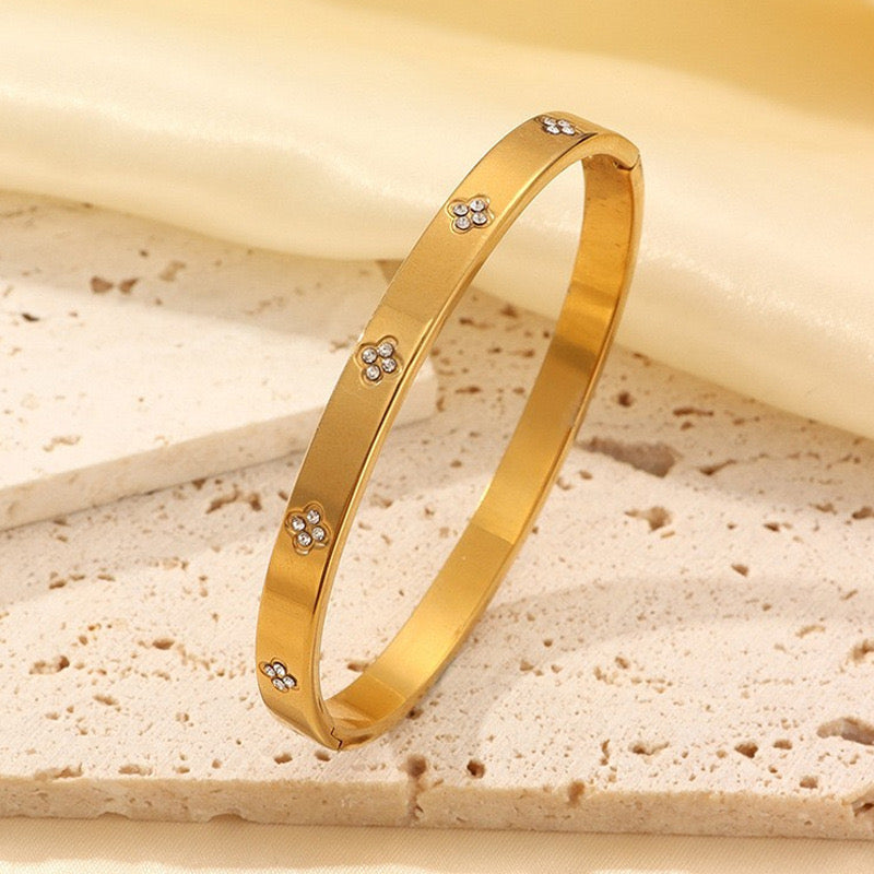 A polished gold bangle bracelet with small diamond-like floral accents, displayed on a textured beige stone surface with a soft cream fabric backdrop.