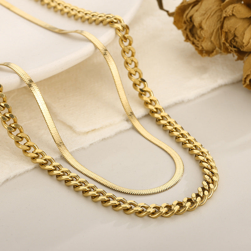 Two gold-tone chains, one thick curb link and one fine snake link, draped on a light beige fabric surface with a dried rose prop in the corner.
